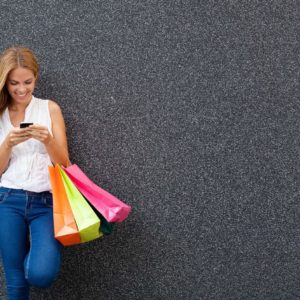 A young woman leaning against a gray textured wall, smiling as she looks at her smartphone, holding multiple colorful shopping bags in her other hand.