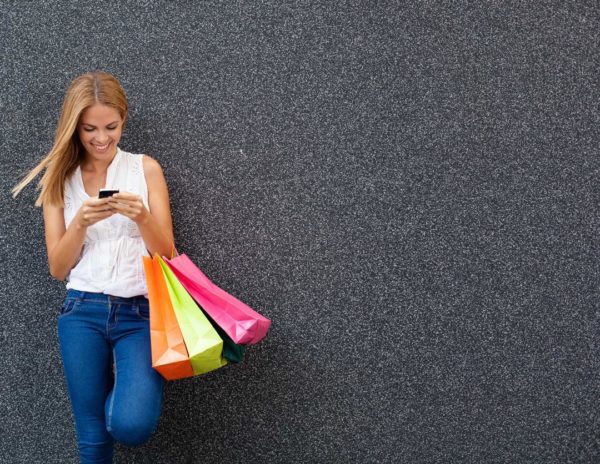 A young woman leaning against a gray textured wall, smiling as she looks at her smartphone, holding multiple colorful shopping bags in her other hand.